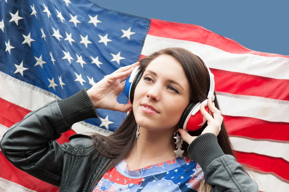 Mujer joven con auriculares escuchando música, con una bandera estadounidense de fondo.