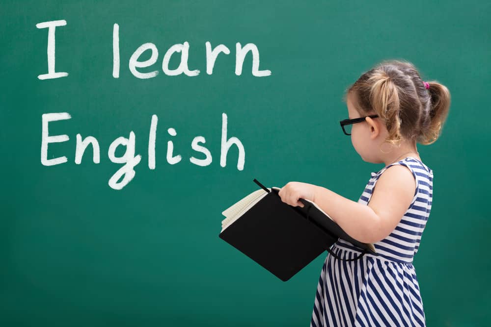 Niña pequeña con gafas leyendo un libro frente a una pizarra verde donde se lee "I learn English". 
