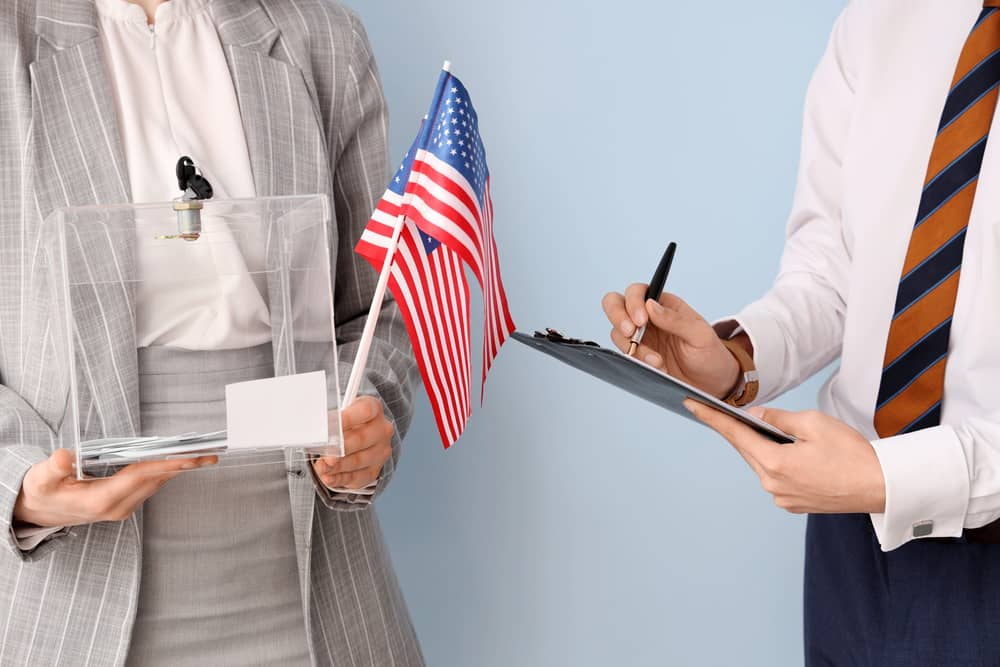 Dos personas de negocios, una sosteniendo una bandera de Estados Unidos y una caja transparente, la otra escribiendo en un portapapeles, simbolizando la etiqueta empresarial en EE.UU.