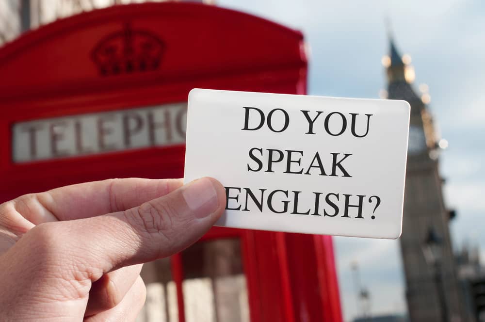 Mano sosteniendo una tarjeta con la pregunta "¿Hablas inglés?" delante de una cabina telefónica roja de Londres y el Big Ben desenfocados al fondo. Ilustración sobre aprender inglés en el Reino Unido.