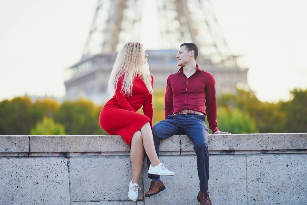 Pareja joven y atractiva sentada en un muro en París, con la Torre Eiffel al fondo, mirándose con afecto y coqueteando. Ella lleva un vestido rojo y él una camisa roja.