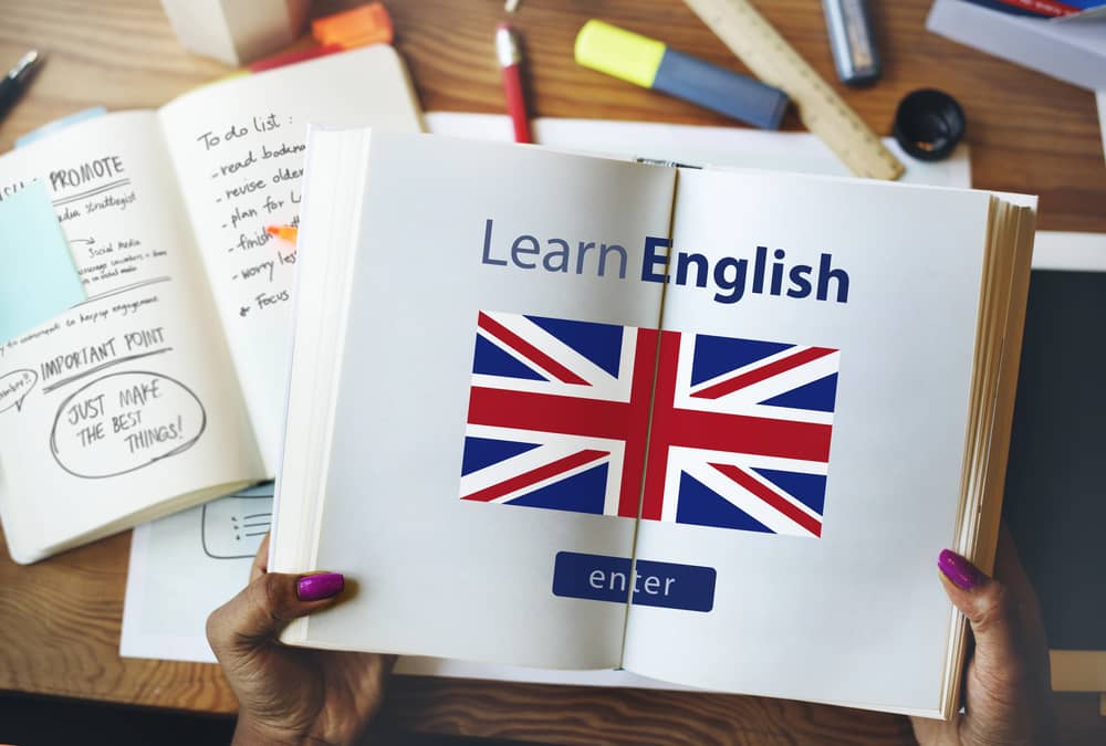 Manos sosteniendo un libro abierto con la portada "Learn English" y la bandera del Reino Unido, junto a un cuaderno con notas de estudio.