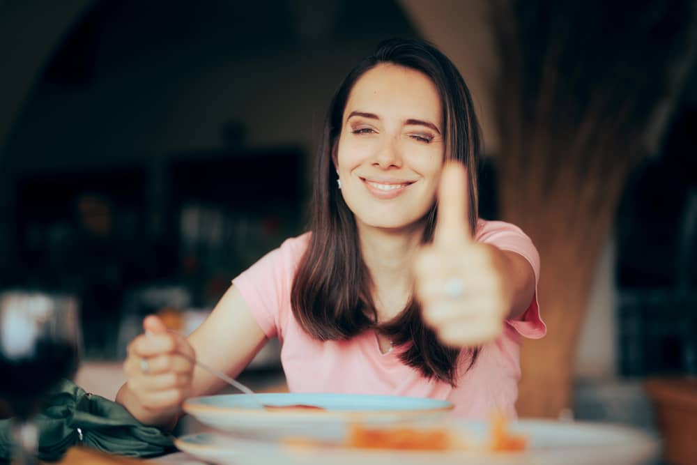 Una mujer sonriente en un restaurante con el pulgar hacia arriba, disfrutando de su comida, representando el éxito al usar frases en inglés para restaurantes.