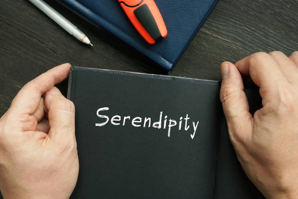 Manos sosteniendo un cuaderno abierto de color negro con la palabra "Serendipity" escrita en blanco, sobre una mesa de madera oscura con un bolígrafo y un marcador.