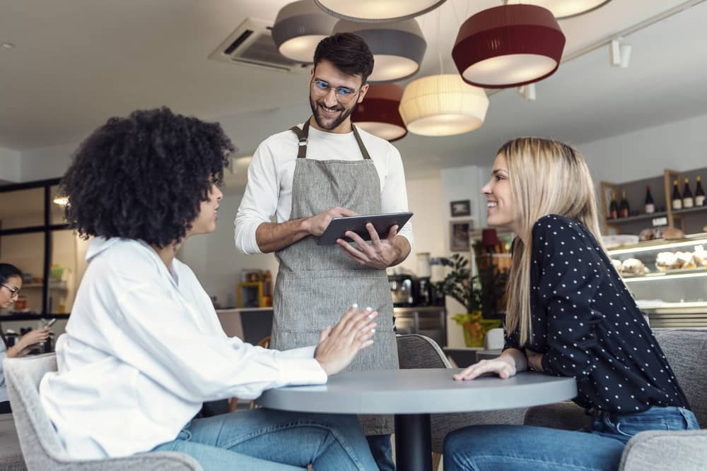 Dos mujeres en una cafetería practican un diálogo en un restaurante en inglés mientras el camarero toma su orden.